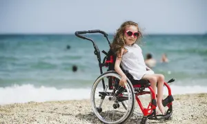 Dziewczynka w wózku inwalidzkim, w białej sukience i okularach przeciwsłonecznych, siedzi na plaży. W tle widać morze i ludzi kąpiących się w wodzie.